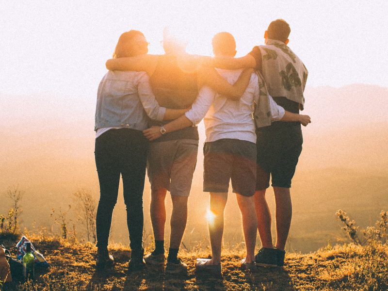 Cuatro amigos de pie en una colina al atardecer, abrazándose mientras miran el horizonte.