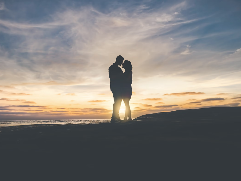 Una pareja abrazada en la playa mientras contemplan un hermoso atardecer junto al mar, con colores cálidos y suaves.