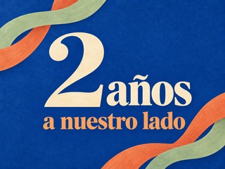 Ilustración de letras crema y albaricoque con cintas de colores sobre azul cobalto, con el mensaje «2 años a nuestro lado»