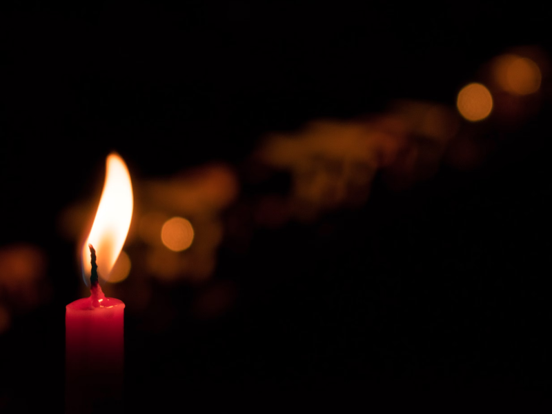 Una vela encendida en la oscuridad, con un resplandor suave y luces desenfocadas al fondo, creando un ambiente cálido y reconfortante.