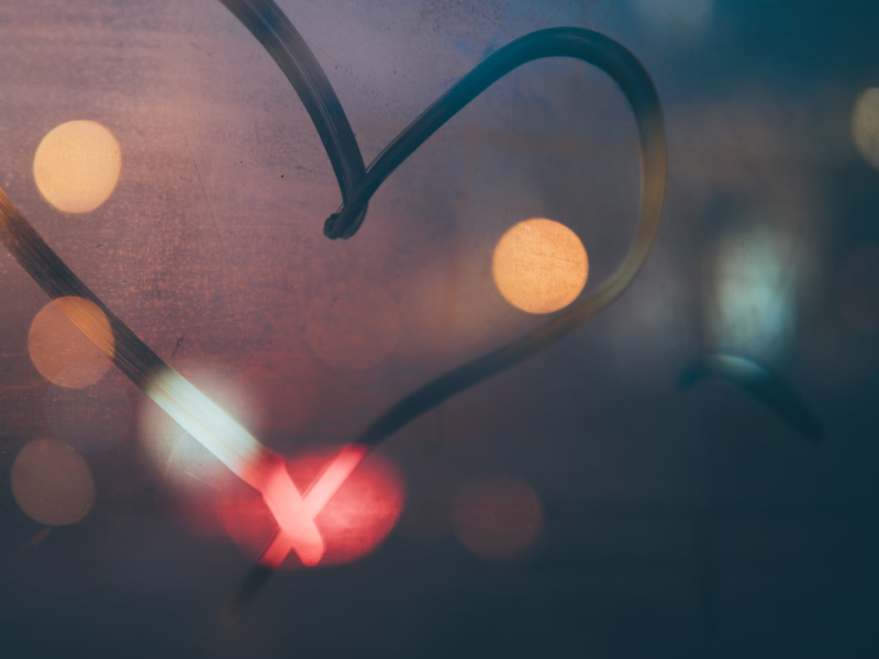 Corazón dibujado en un vidrio empañado con luces desenfocadas al fondo.